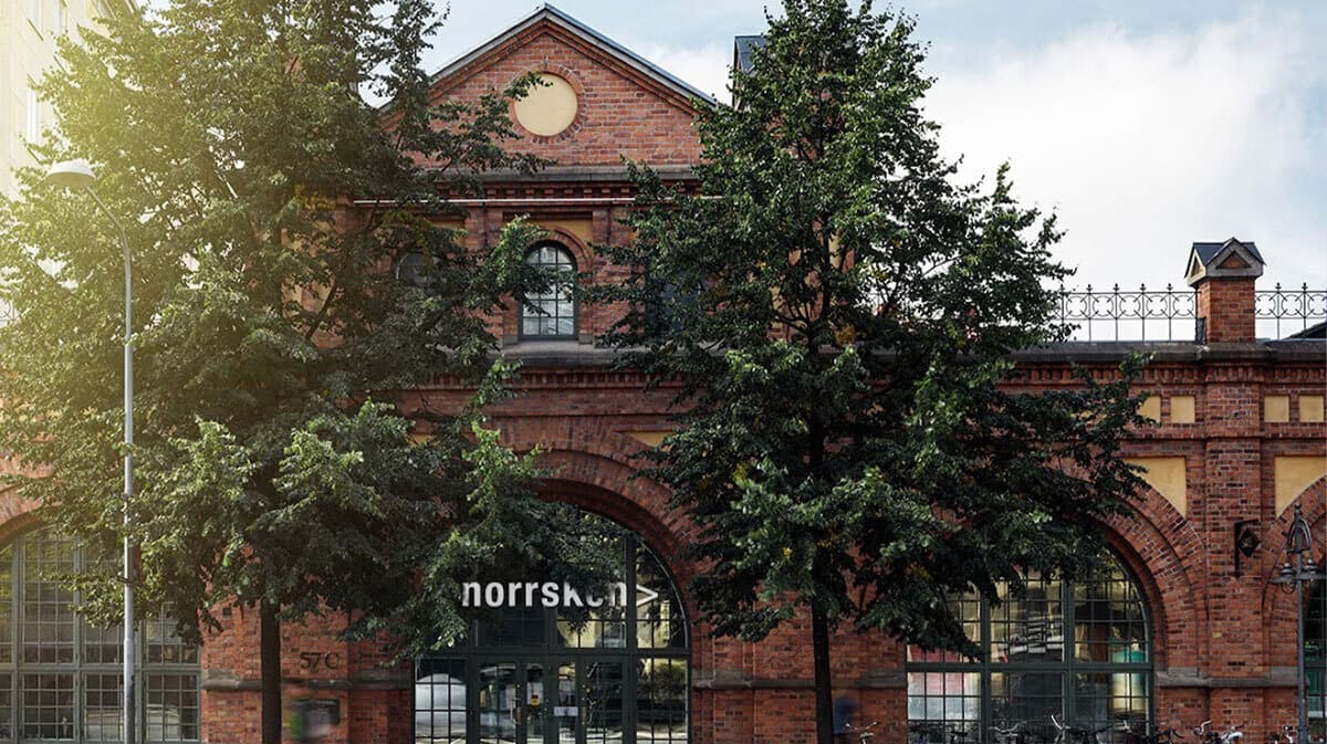 Photo of the red-brick Norrsken House building in Stockholm, its arched entrance and "norrsken" signage framed by two trees.