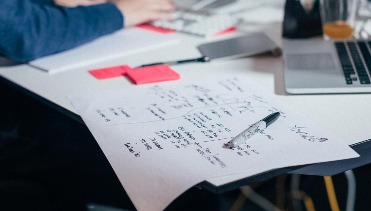 A sheet of handwritten notes and arrows on a desk, a pen resting on the paper between laptop and sticky notes.