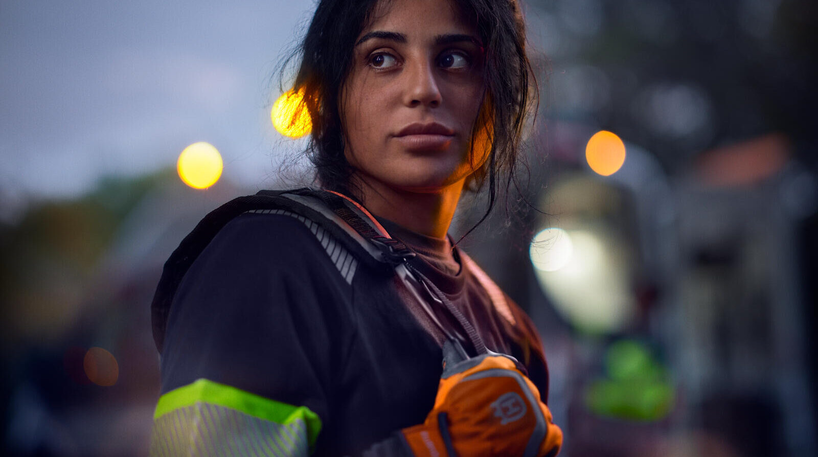 Night portrait of a young woman in reflective workwear, street lights blurring to warm bokeh behind her.