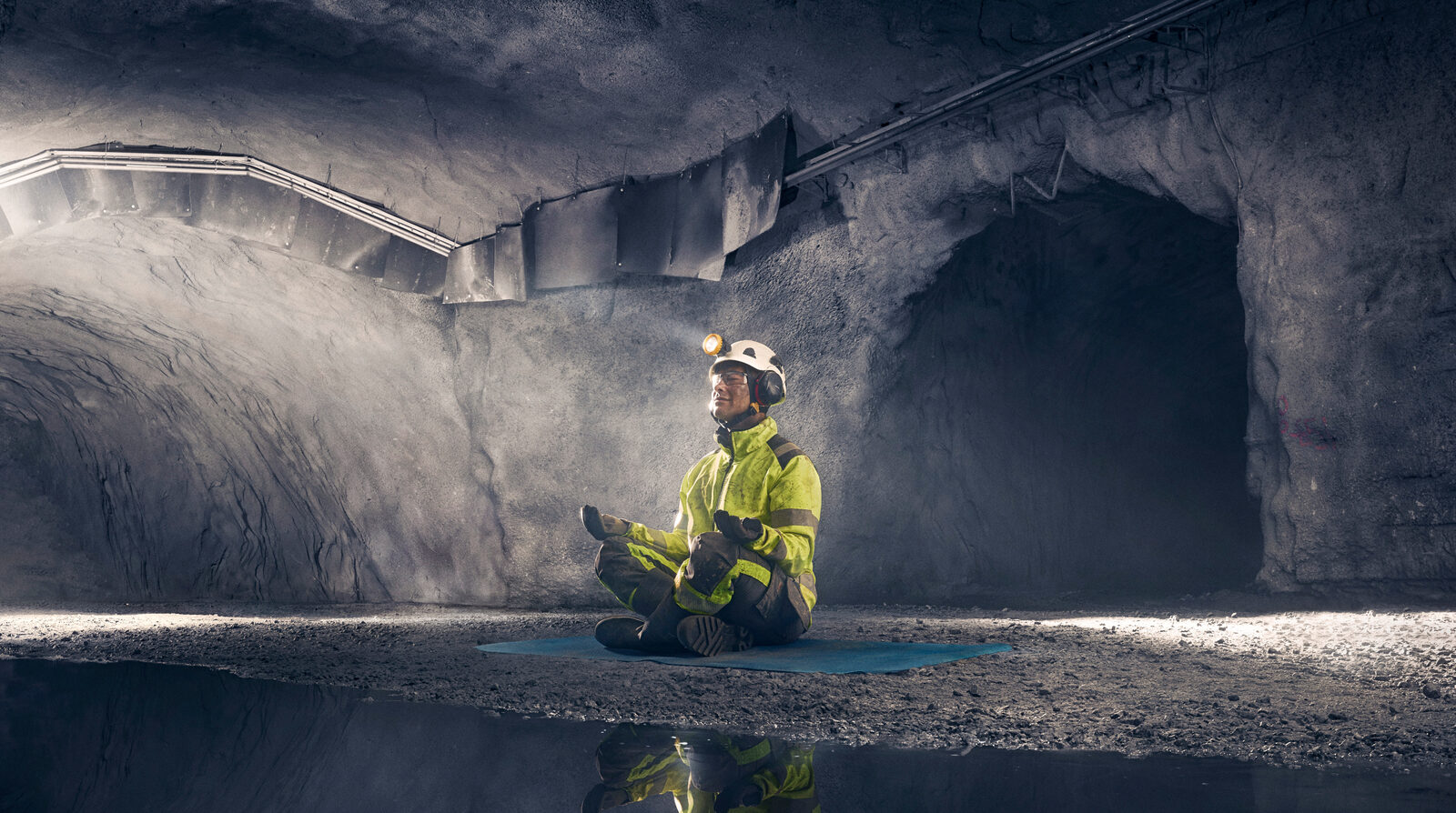A worker in hi-vis and helmet sits cross-legged in an underground mine tunnel, meditating above a mirror-still pool.