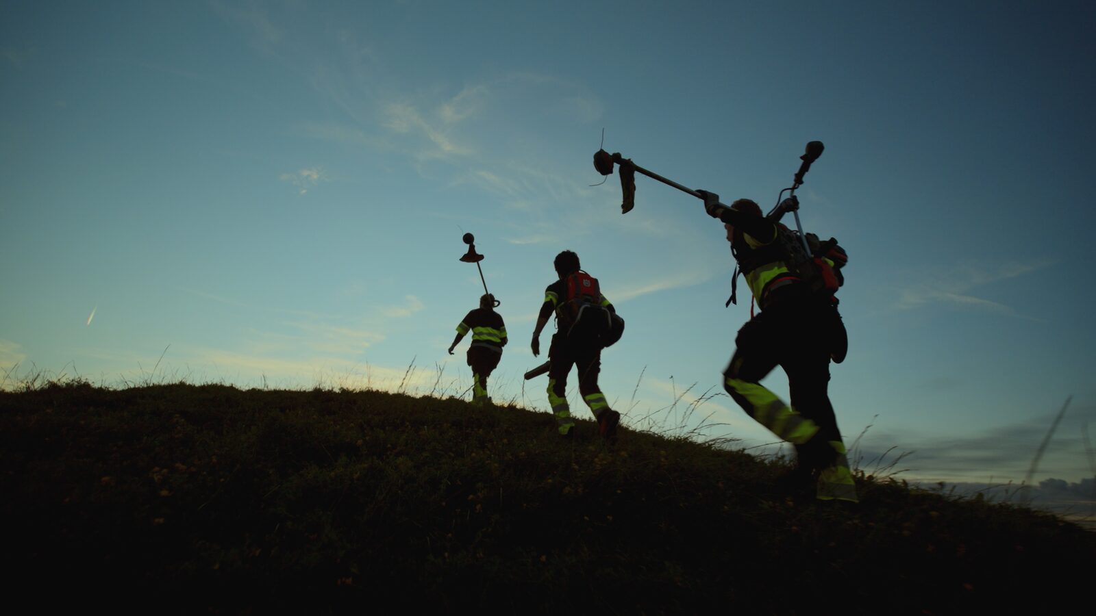 A crew in hi-vis silhouetted against the dusk sky, carrying gear up a grassy hillside.