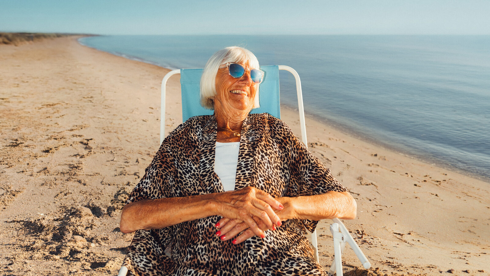 An older woman in blue sunglasses and leopard print grins from a beach chair by the sea.
