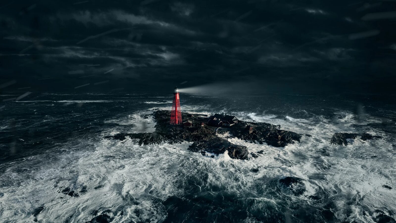 A small red lighthouse beams through a storm as waves break white over dark rocks.