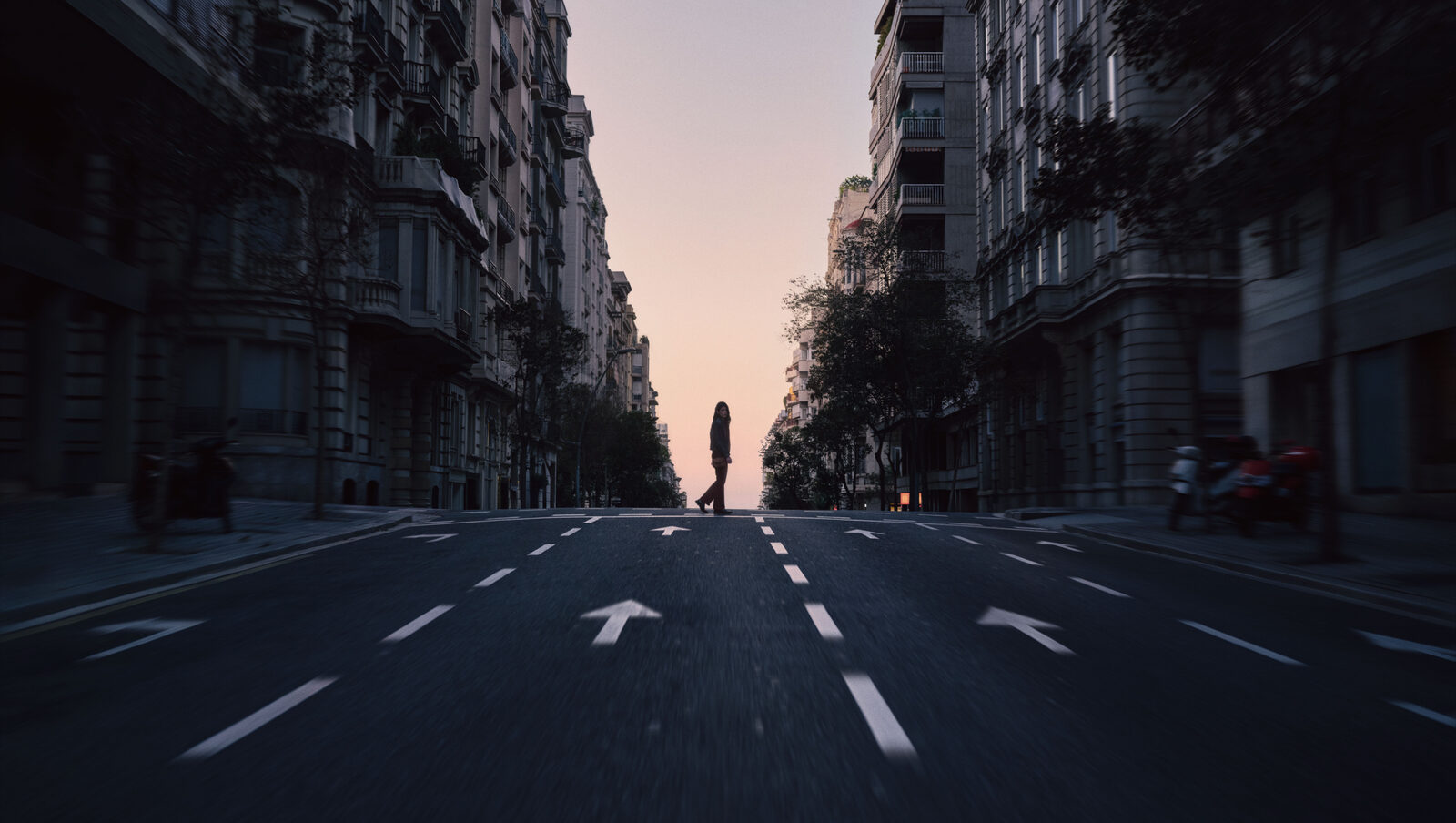 A lone figure stands far down an empty city street at dusk, lane arrows pointing the way ahead.