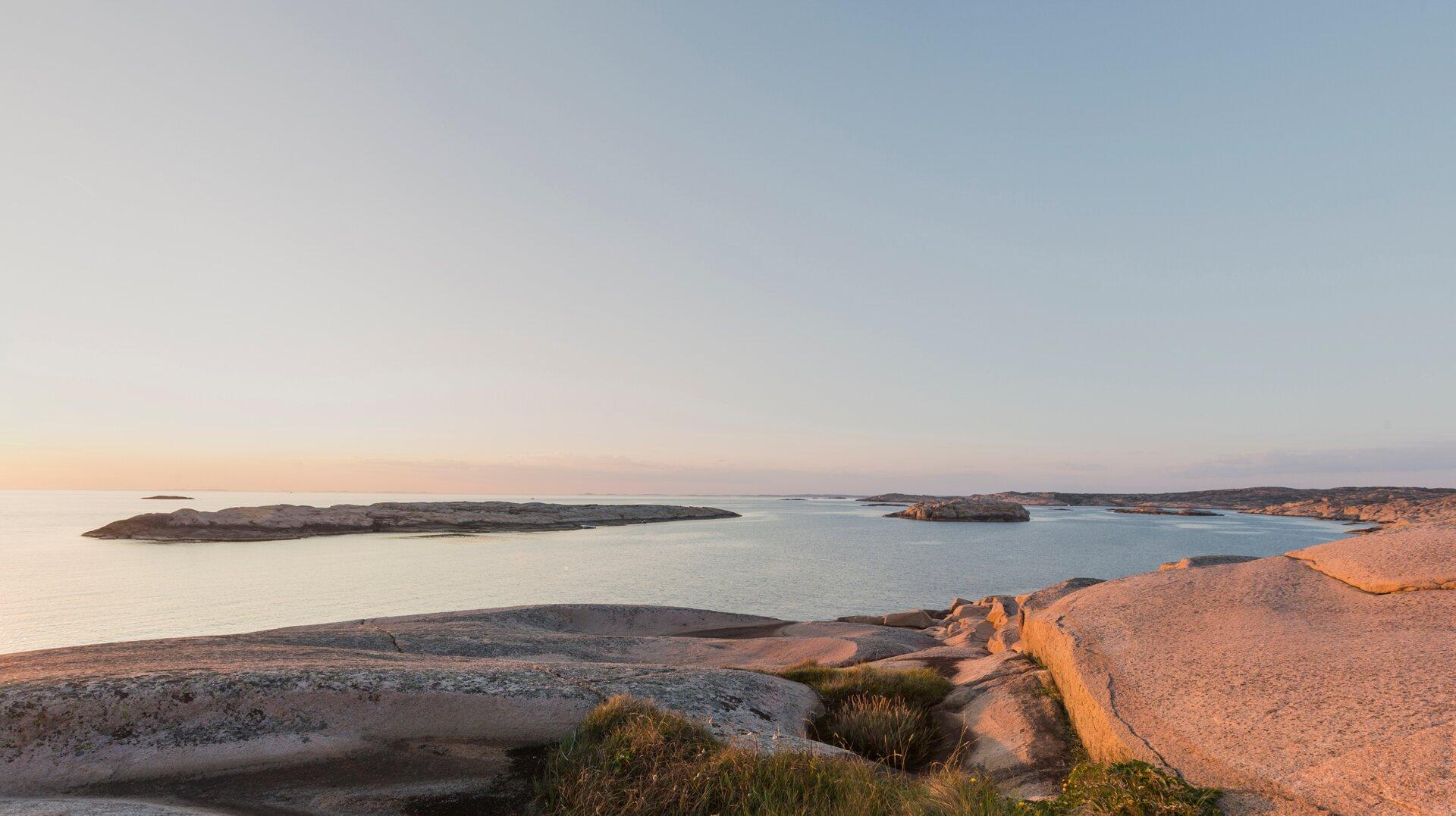 Smooth granite rocks slope down to calm archipelago water at sunset, low islets dotting the horizon under a clear sky.