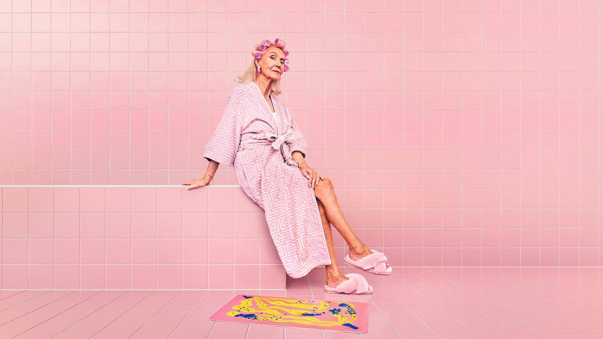 An older woman in a pink bathrobe and hair curlers reclines on the edge of a pink-tiled bathroom, fluffy pink slippers beside a yellow-and-blue patterned anti-slip mat – the campaign key visual.