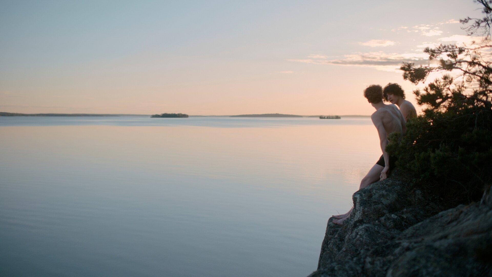 Two young people sit close together on a rocky outcrop at dusk, looking out over a still sea at sunset – a quiet moment from the För all tid film.