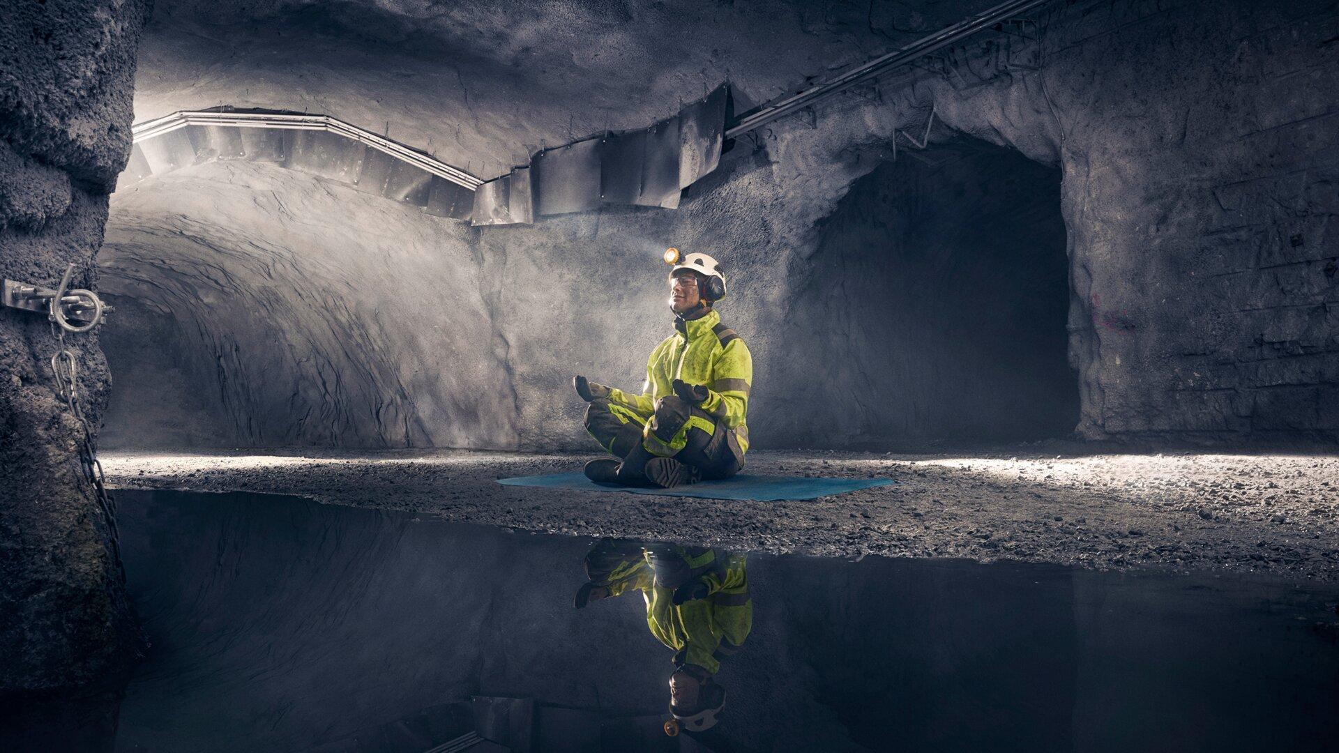 A worker in hi-vis jacket and helmet sits cross-legged on a mat, meditating in an underground mine tunnel, mirrored in still water.