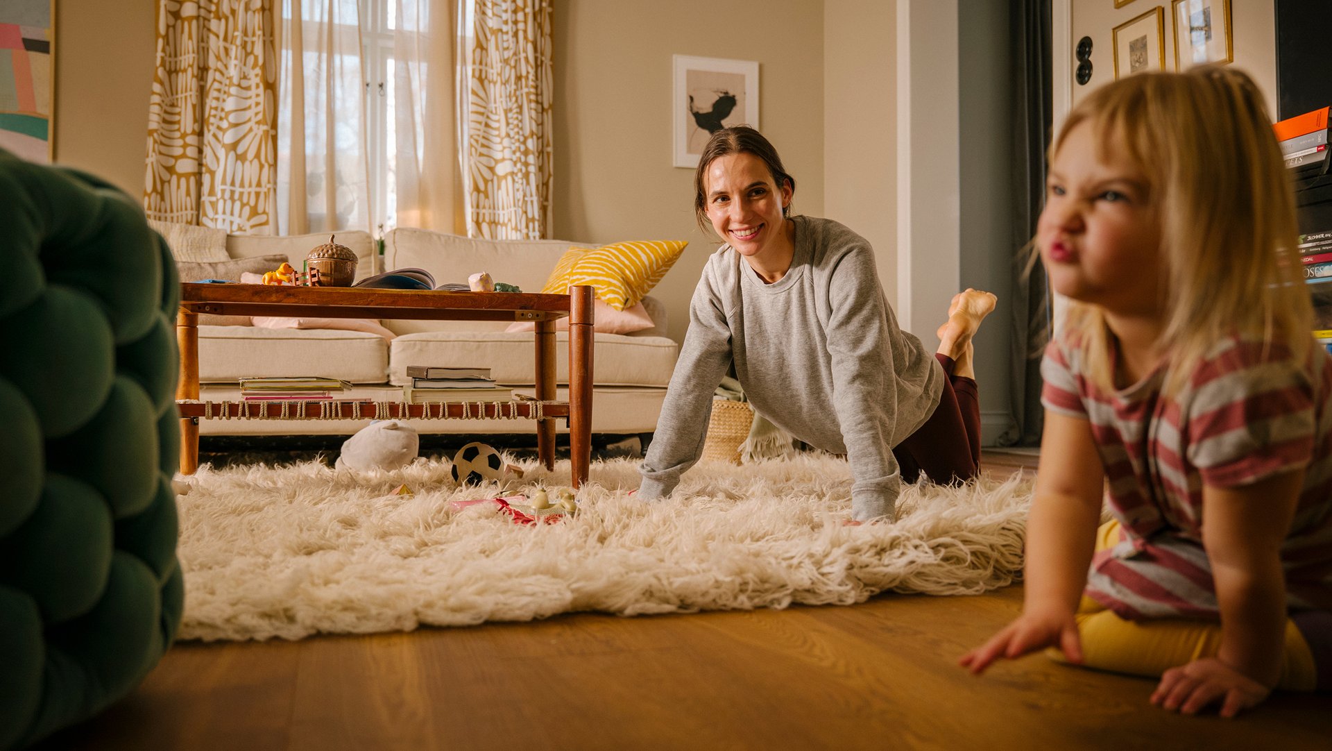 A film still of a mother doing push-ups on the living-room rug while her daughter plays beside her – everyday exercise, no gym required.
