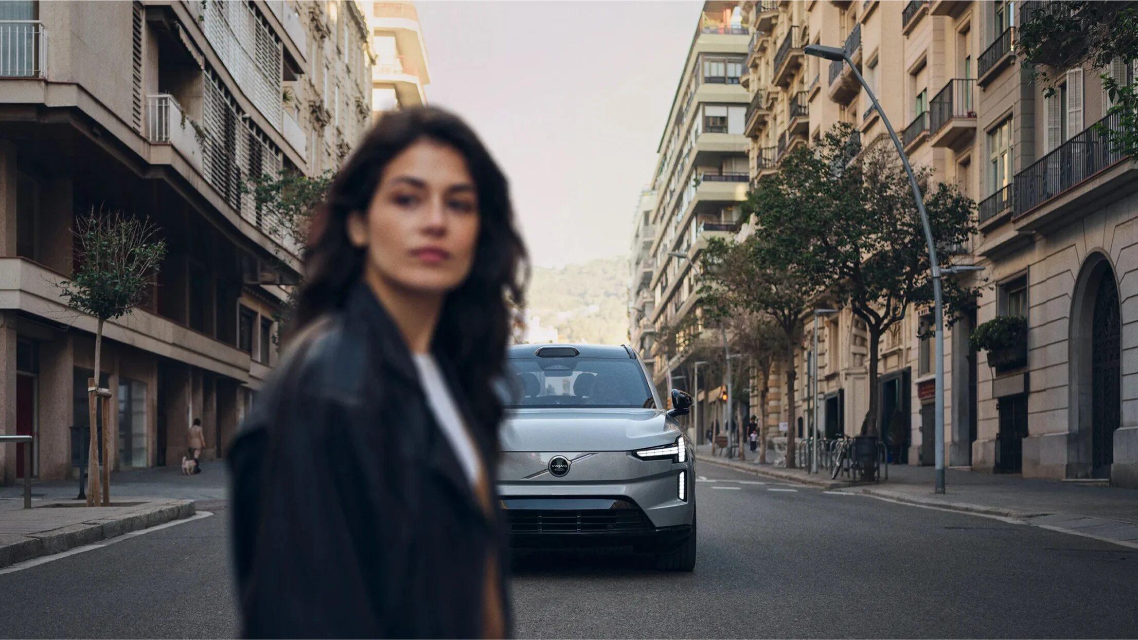A woman in a black leather jacket on a quiet city street, a silver electric SUV in sharp focus behind her.