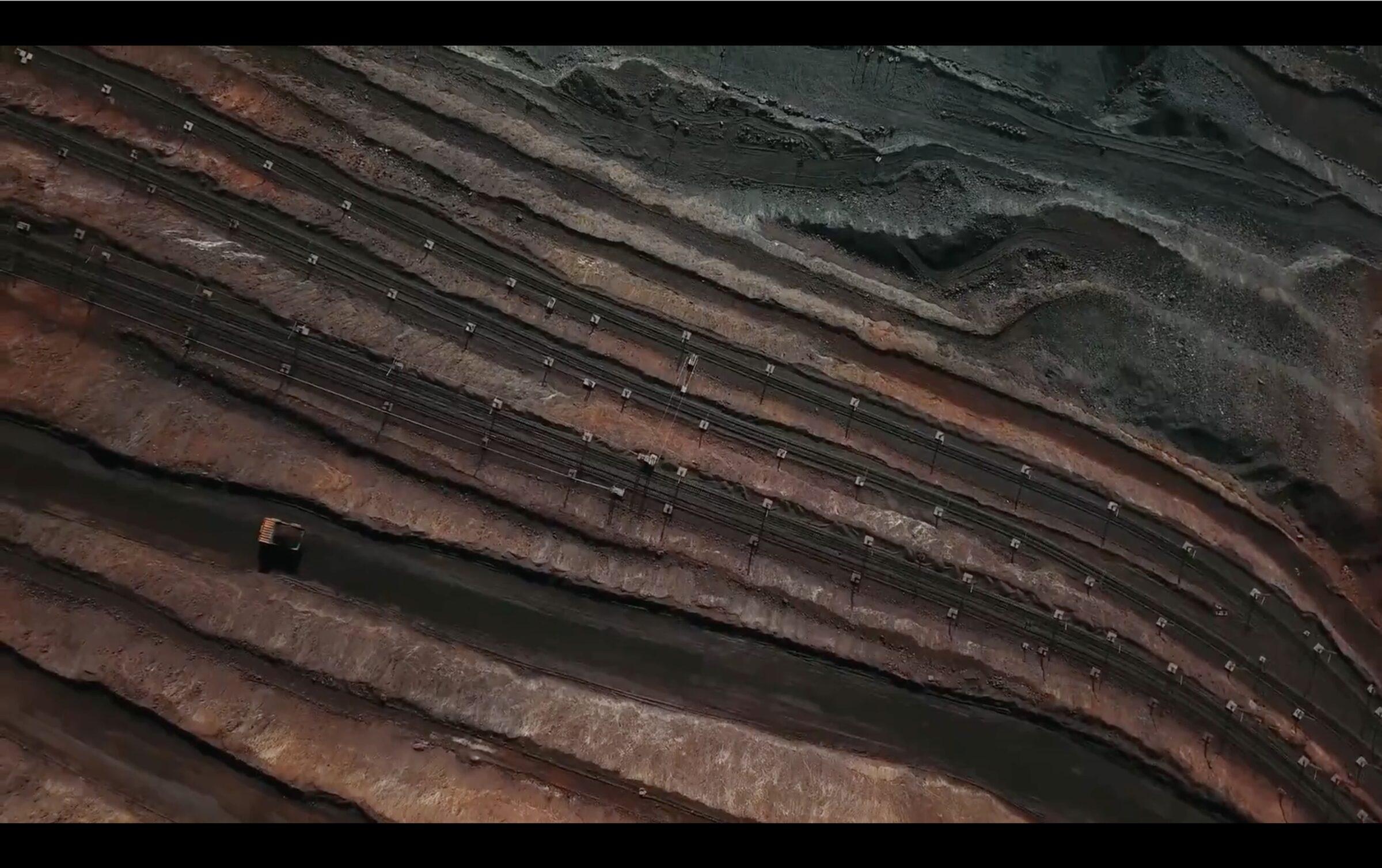 Aerial view of a terraced open-pit mine, red-brown rock cut into bold diagonal layers.